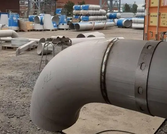 Ulkona on suuri harmaa putki, jossa on metallinen kiinnike, ja taustalla on muita putkia ja rakennus.