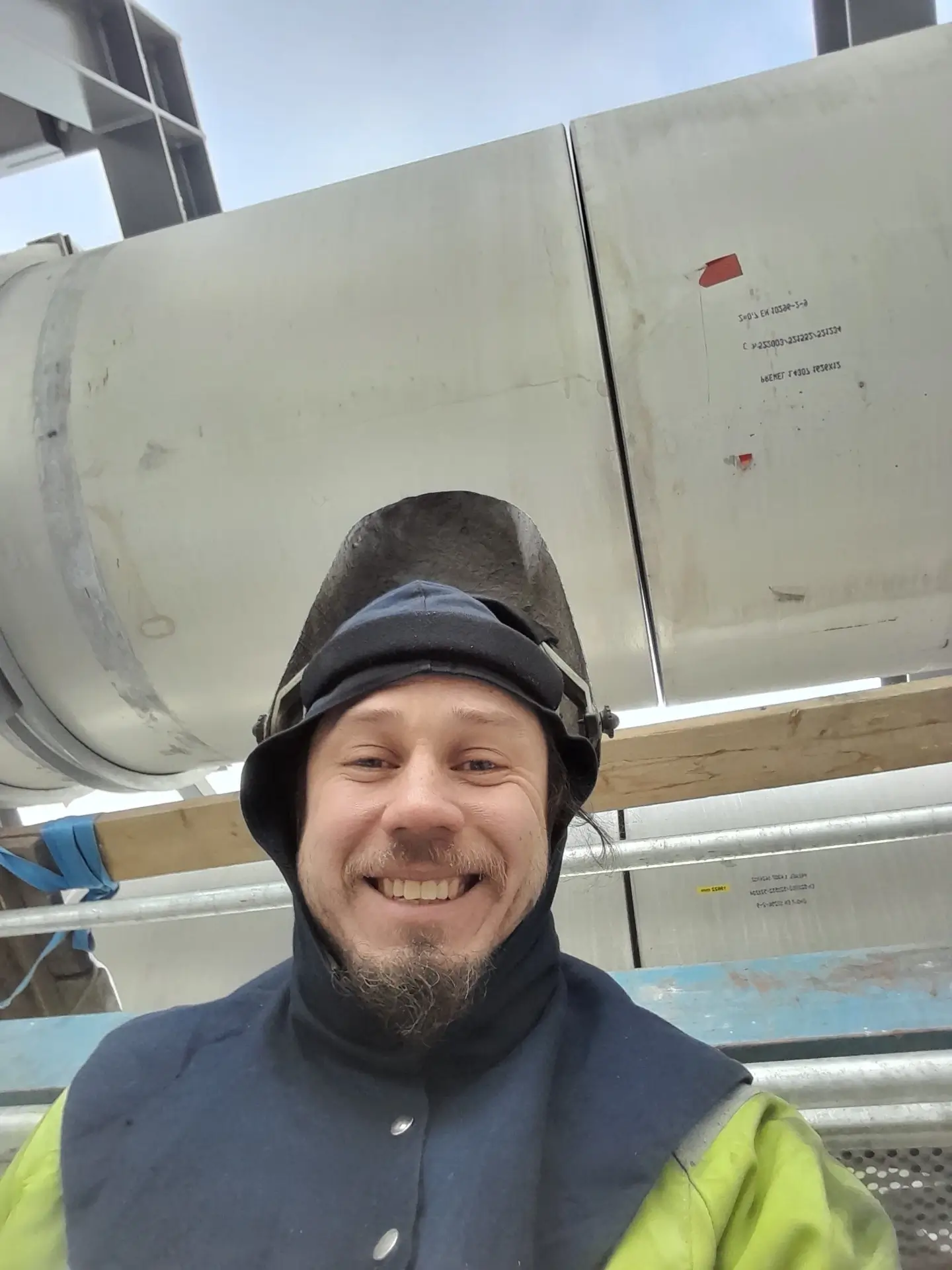 Hymyilevä mies hitsausmaski päässä, taustalla metalliputkia ja rakennustelineitä, valoisa ja aurinkoinen sää.