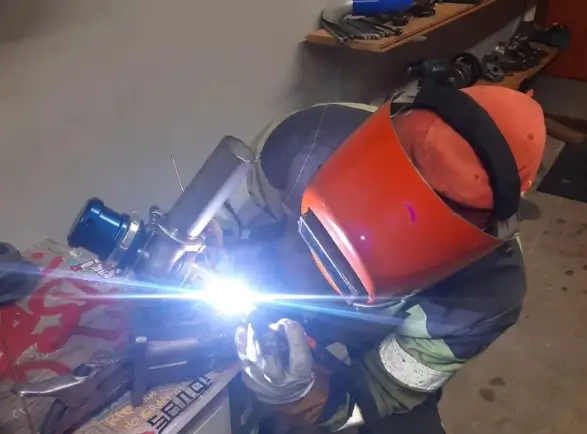 Hitsaaja hitsaa metallia suojavarusteissa, kirkas valo, työkaluja hyllyllä.