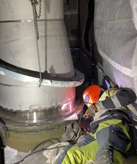 Hitsaaja työskentelee metallirakenteen parissa, suojavarusteet päällä, kirkkaat valokaaret valaisevat työtä.