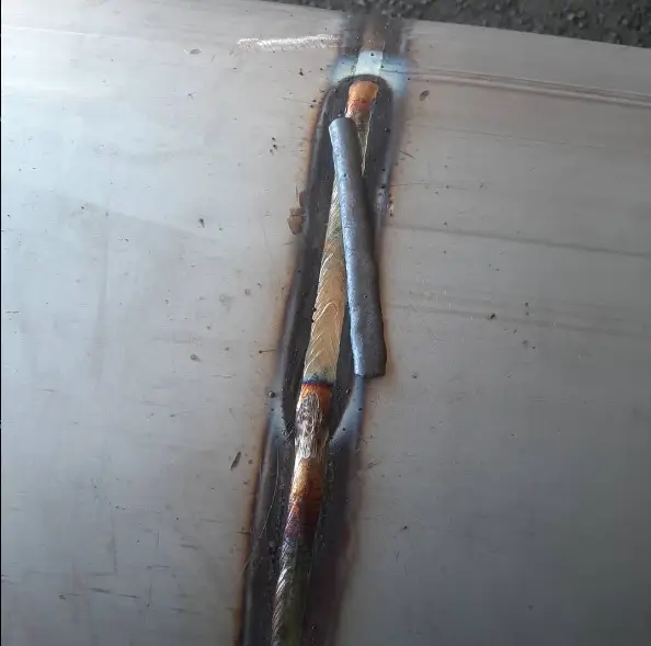 Metallipinnalla on hitsausjälki, jossa näkyy ruskeita ja harmaita sävyjä, sekä metallitanko. Valo on kirkas ja tasainen.