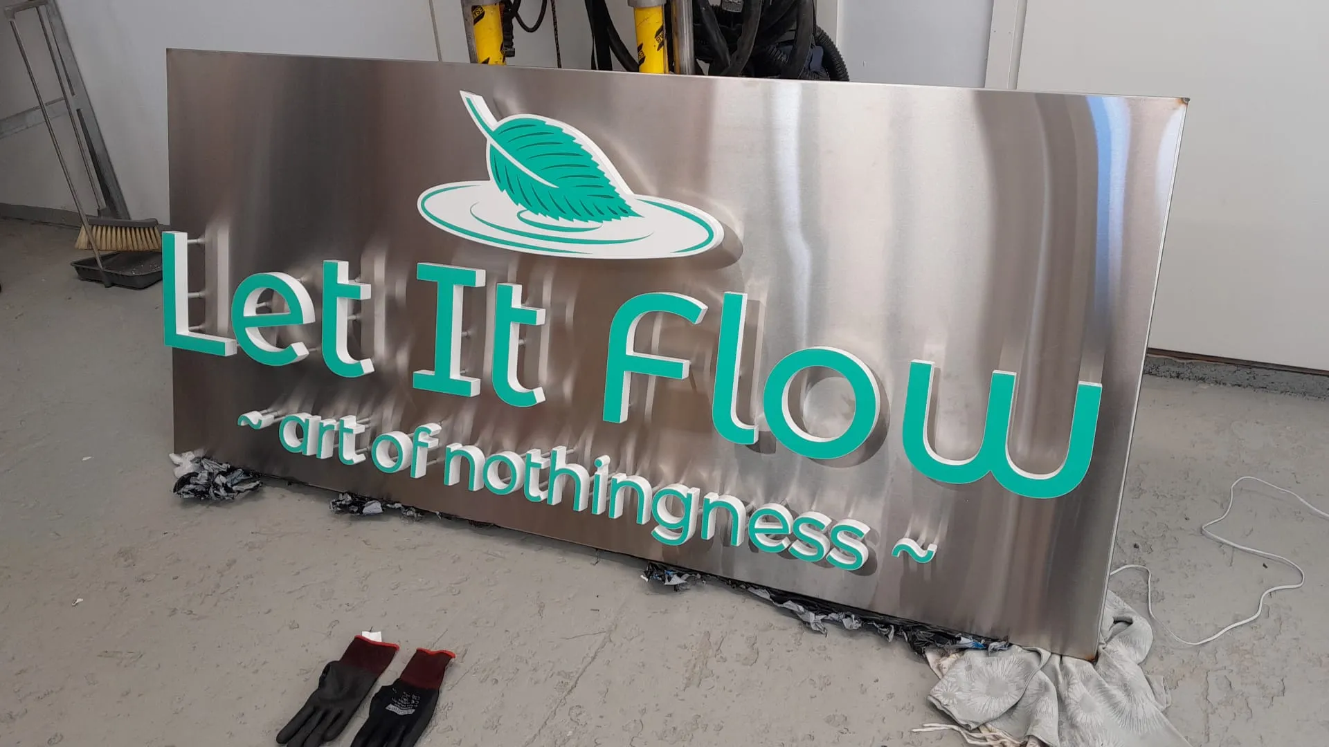 Suuri metallinen kyltti, jossa on kohokirjaimet 'Let It Flow' ja lehtilogo, makaa teollisuuden työtilan lattialla.