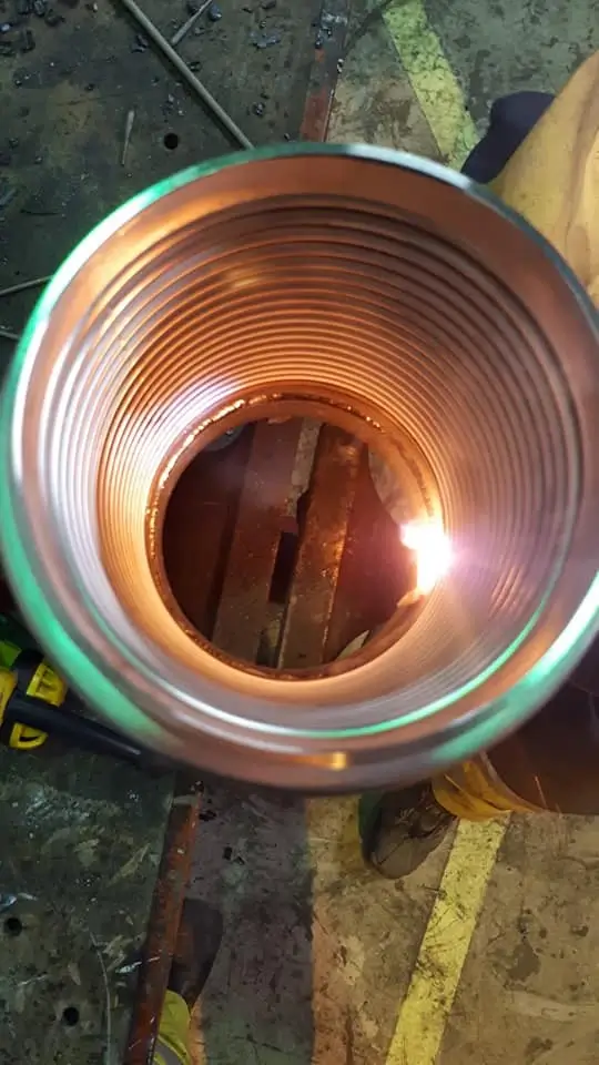 Metallinen putki, jossa on sisäkierteet ja hitsausliekki keskellä, valaistu lämpimällä valolla.