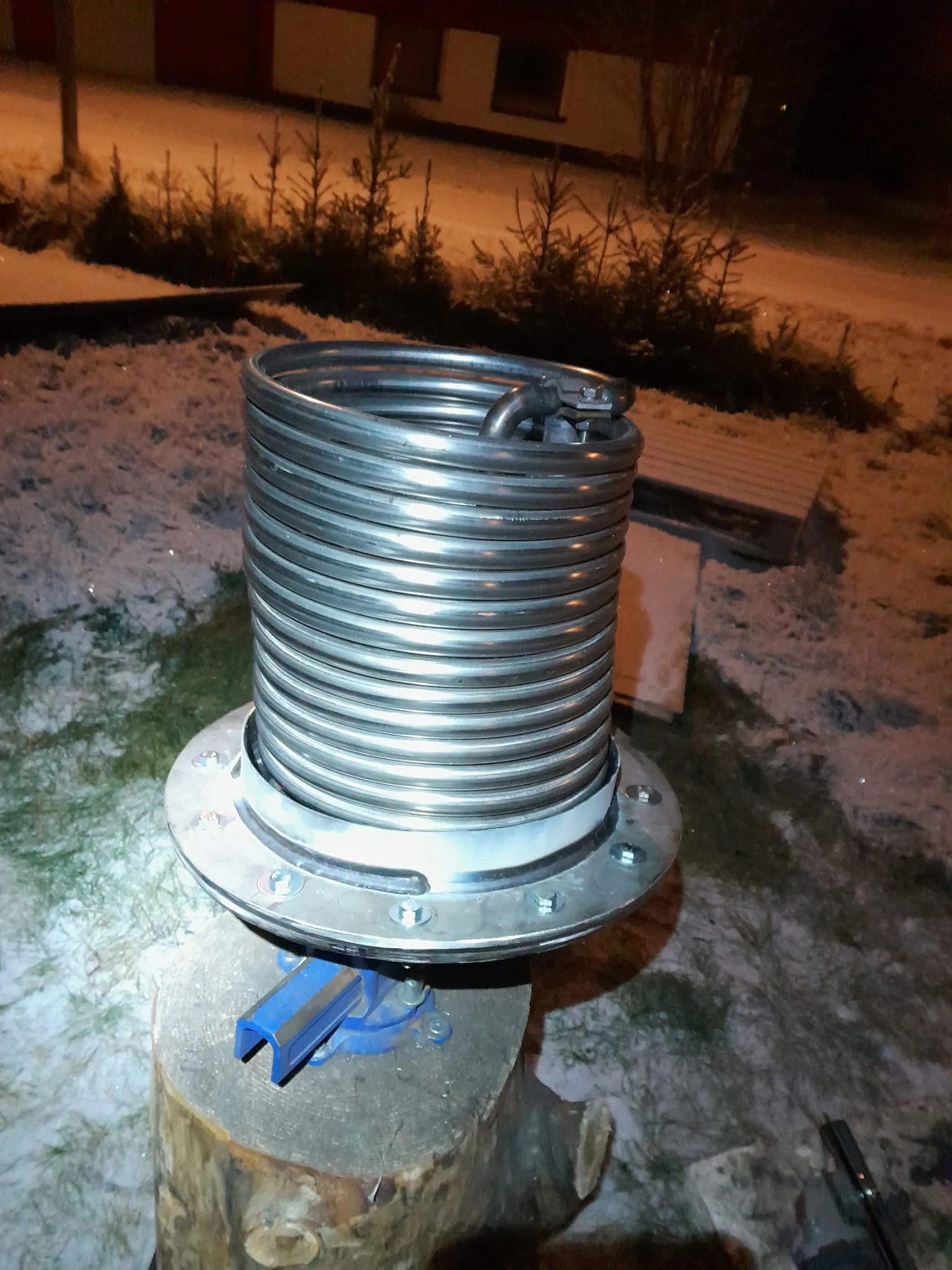 Metallinen spiraali, jossa on laippa, on kiinnitettynä siniseen ruuvipuristimeen puupölkyllä ulkona, jossa on lunta ja puita taustalla.