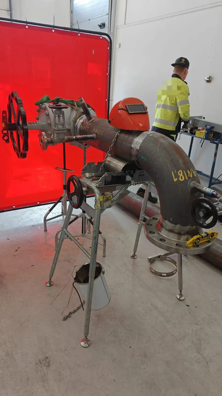 Kuvassa on metallinen putkisto, jossa on venttiilejä ja laippoja, asetettuna metallisille telineille punaisen hitsaussuojan edessä. Oranssi hitsausmaski on putken päällä. Taustalla on henkilö keltaisessa takissa ja työpöytä.
