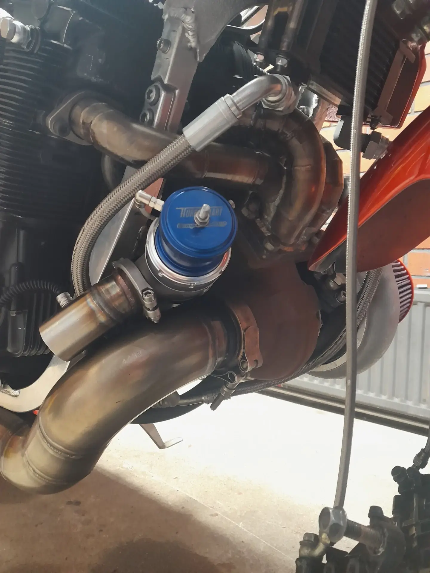 Lähikuva moottoripyörän moottorista, jossa näkyy turboahdin, pakoputket ja sininen venttiili, metallisilla letkuilla ja kirkkaalla valaistuksella.