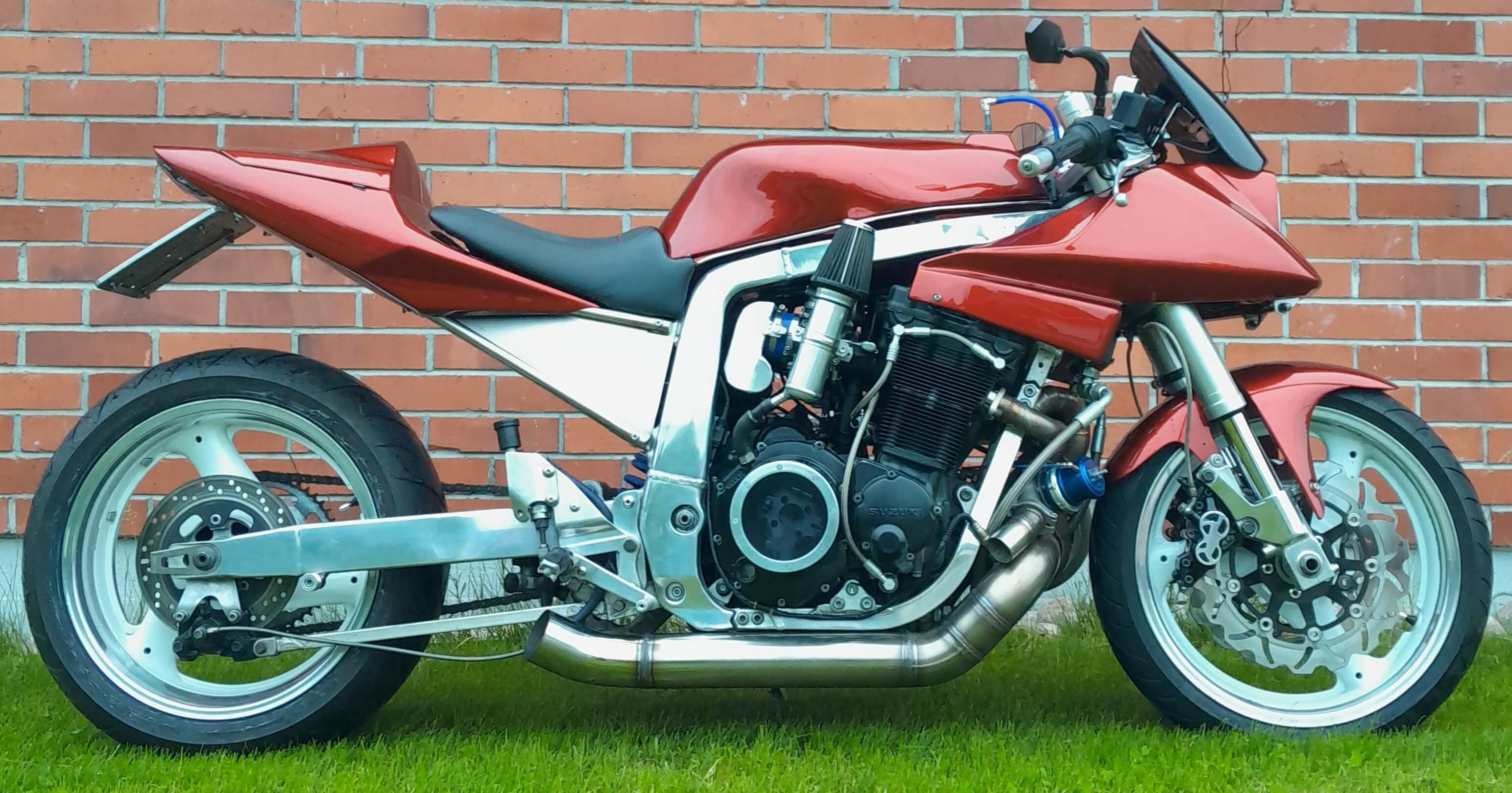 Punainen moottoripyörä seisoo vihreällä nurmikolla tiiliseinää vasten, jossa on hopeanvärisiä yksityiskohtia ja musta istuin.