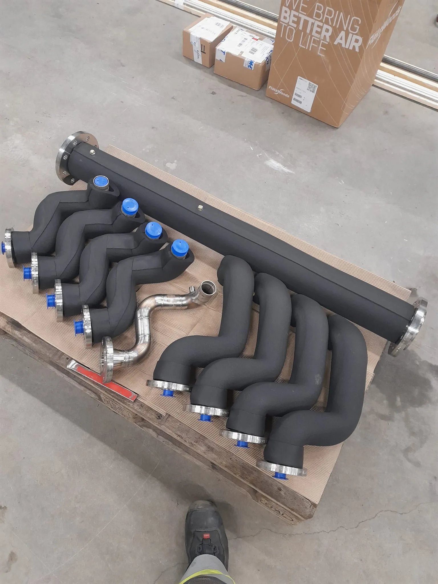 Kuvassa on puulaatikko, jossa on mustia putkia, joissa on sinisiä korkkeja ja metallisia laippoja, sekä yksi kiiltävä metalliputki. Taustalla on pahvilaatikoita ja metallitankoja.