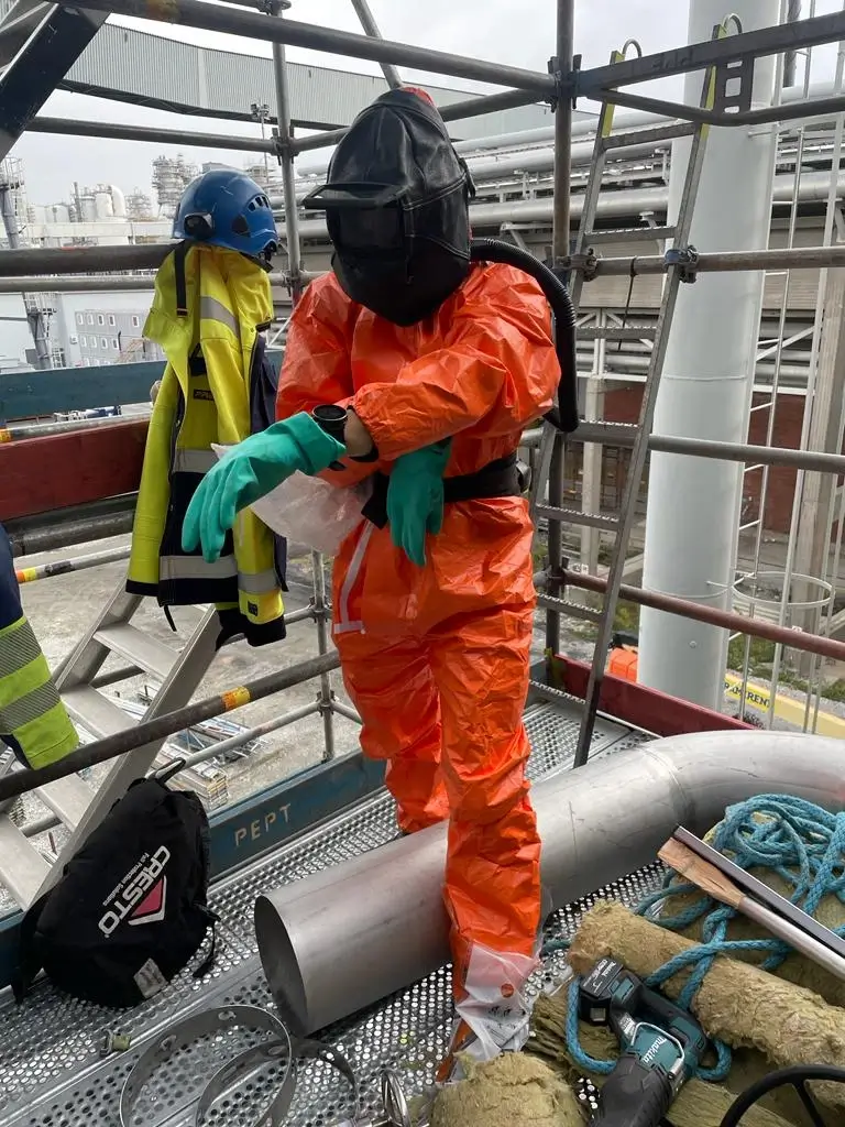 Oranssiin suojapukuun pukeutunut henkilö seisoo rakennustelineillä, yllään musta suojakypärä ja vihreät käsineet, taustalla teollisuuslaitos ja työkaluja.