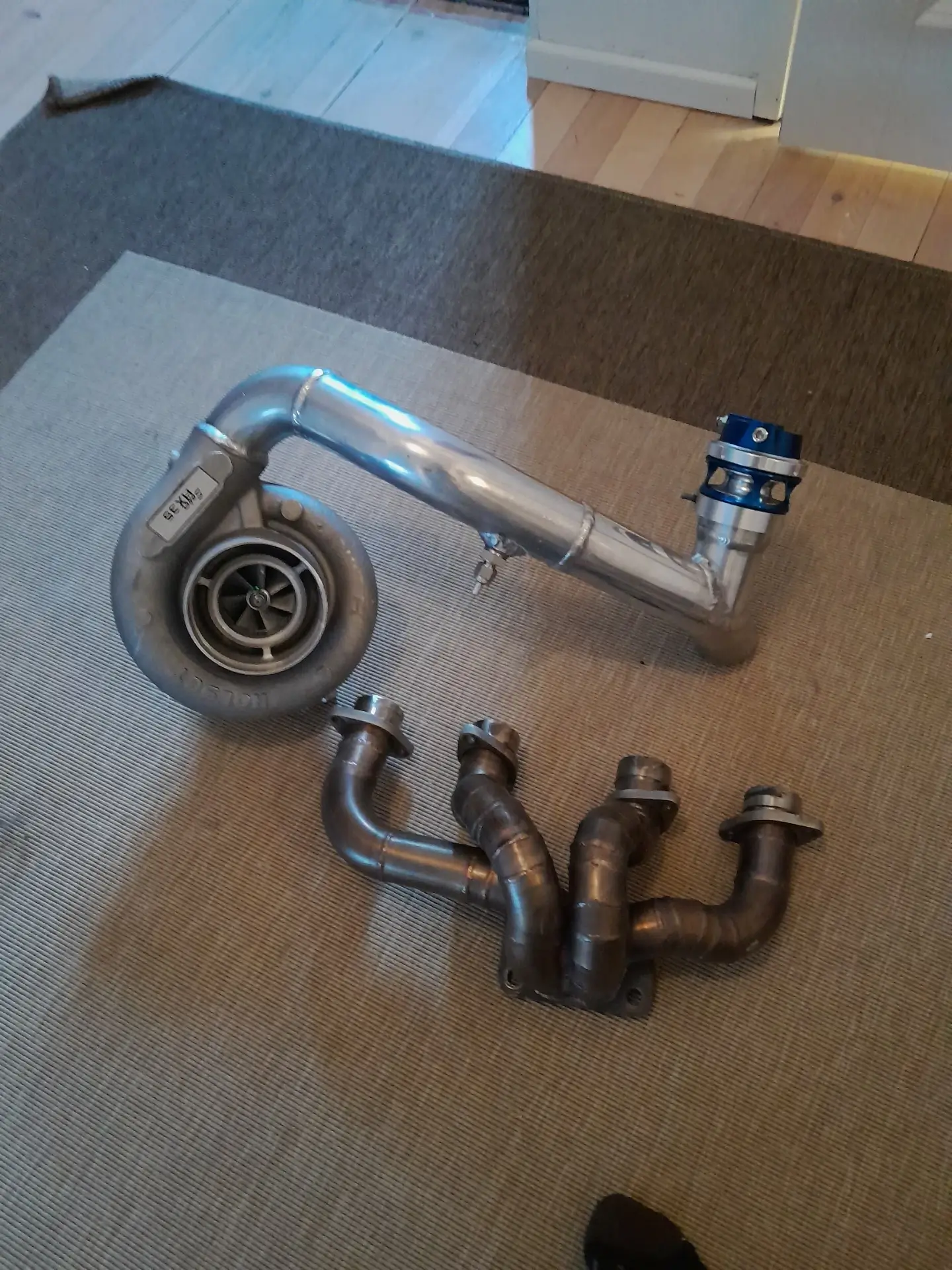 Kuvassa näkyy turboahdin, pakosarja ja hukkaportti harmaalla matolla. Metalliset osat ovat hopeanvärisiä ja sinisiä. Valo on kirkas.